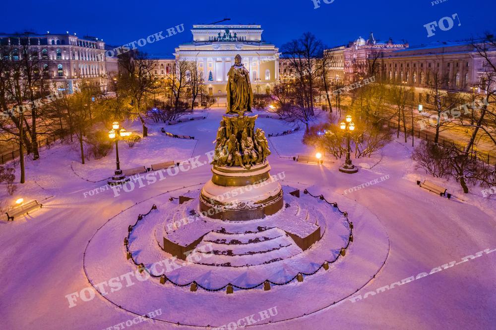 На фотографии изображен зимний вечер в Санкт-Петербурге. Показана площадь с памятником Катерине Великой, окружённым снежным парком и фонарями, создающими уютную атмосферу. На заднем плане виден Александринский театр, его фасад освещён. Это изображение подходит для использования в туристических проспектах, статьях о зимних праздниках в России, иллюстрациях о культурном наследии Санкт-Петербурга, а также может использоваться для оформления открыток или календарей.